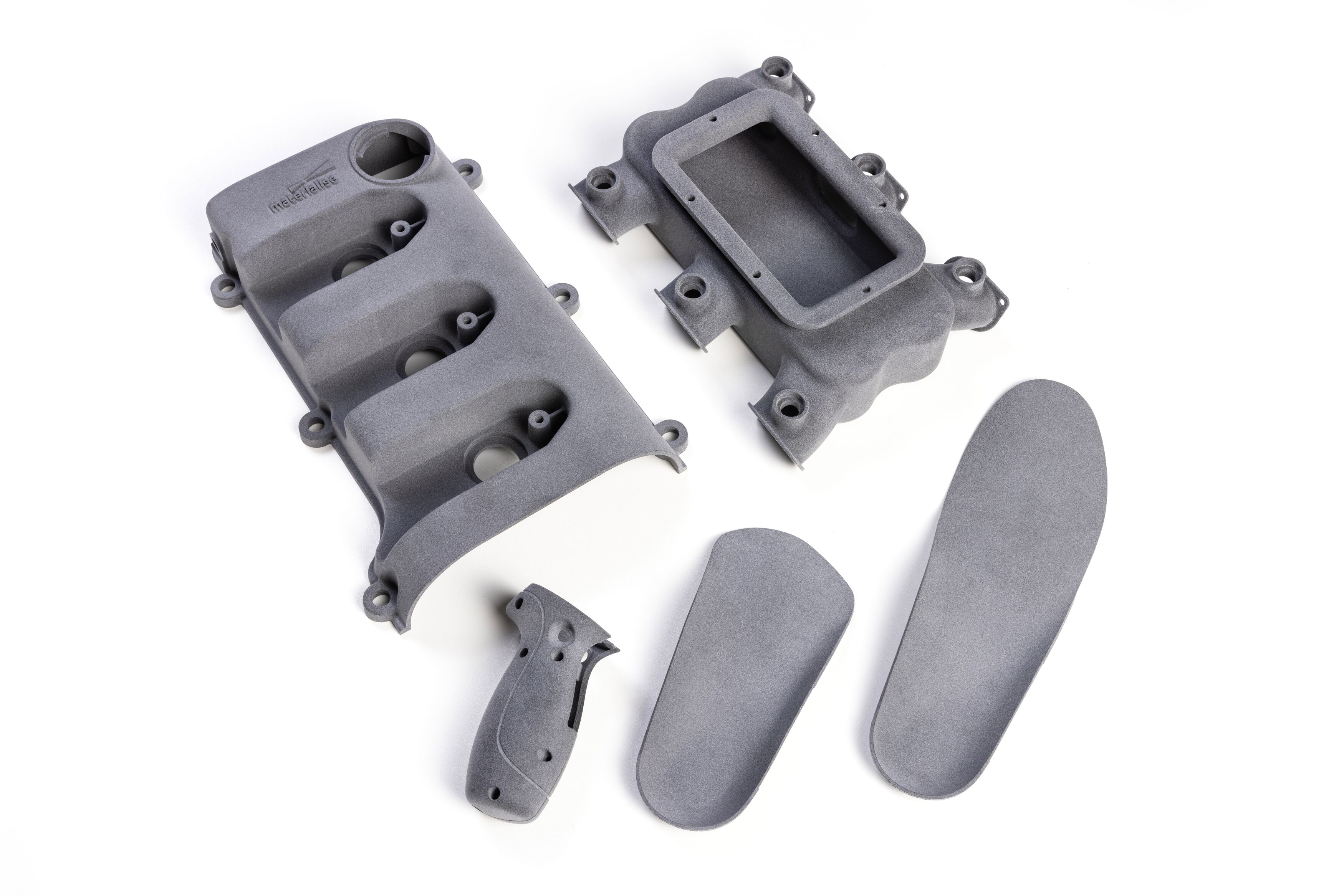 A collection of grey 3D-printed parts made in Polyamide 11 using MJF, including orthotic insoles, a handle, and two pieces of housing.